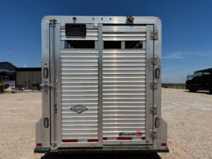 A Frontier 20' Stock Combo livestock trailer is shown from the rear, featuring horizontal slats, two latch handles, and a Winchester logo. The trailer sits on gravel with buildings and vehicles in the background under a clear blue sky. -Stillwater Trailer Sales
