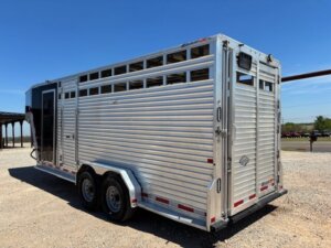 A silver Frontier stock combo livestock trailer, 20' long with ventilation slats and dual axles, is parked on a gravel lot under a clear blue sky. -Stillwater Trailer Sales