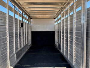 Interior view of an empty Frontier stock combo trailer, 20' long, featuring ribbed aluminum walls, black flooring, and open roof beams with sunlight streaming in from the open back. -Stillwater Trailer Sales