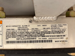 A close-up of a manufacturer&rsquo;s label on a Frontier Stock Combo 20' trailer, displaying specifications such as weight limits, tire pressure, manufacturing date, VIN, and other details in both English and French. The label is attached with two bolts. -Stillwater Trailer Sales