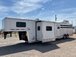 A large white Logan Stock trailer with Combo Living Quarters and a slide-out is parked on gravel under a partly cloudy sky. -Stillwater Trailer Sales