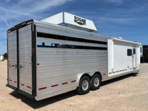 A large silver Logan Stock Combo livestock trailer with slatted sides and dual axles is parked on gravel under a blue sky. The trailer has a white section with windows and sits near a sign that reads Logan Coach. -Stillwater Trailer Sales
