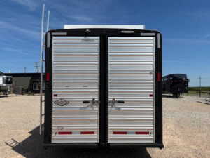 The image shows the rear view of a silver Logan Stock Combo livestock trailer with closed double doors, parked on a gravel lot under a clear blue sky. The trailer has horizontal ridges and black trim. -Stillwater Trailer Sales