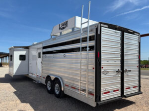 A silver Logan Stock Combo livestock trailer with two axles, rear double doors, side vents, and a roof ladder is parked on gravel under a blue sky. -Stillwater Trailer Sales