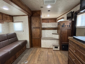 Interior of a Logan Stock Combo RV with wood cabinets, a brown sofa, carpeted steps leading to a small bed, wall-mounted TV, and kitchen area with wood flooring plus neutral-colored walls and window blinds. -Stillwater Trailer Sales