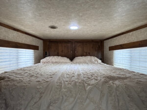 A cozy bed with textured white bedding sits against the back wall of this Logan Stock Combo Living Quarters, flanked by wooden cabinets and two windows with blinds, creating a snug, enclosed sleeping space. -Stillwater Trailer Sales