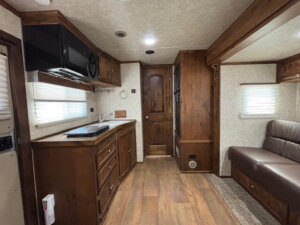 Interior of a Logan Stock Combo Living Quarters camper with wood cabinets, small sink, microwave, countertop, brown door, sofa with brown upholstery, and window blinds. The clean, compact space features a wood-patterned floor for a cozy feel. -Stillwater Trailer Sales