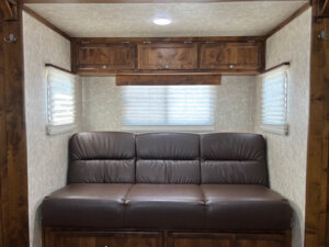 A brown leather sofa sits against the back wall of a Combo Living Quarters space, with wood cabinets above and windows with blinds on either side, creating a cozy, enclosed seating area. -Stillwater Trailer Sales