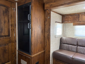 A cozy Logan Stock Combo RV interior with wood-paneled walls, a black double-door refrigerator, a brown leather sofa, two windows with blinds, and built-in wooden cabinets above the seating area. -Stillwater Trailer Sales