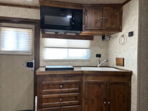 Small RV kitchen in Logan Stock Combo Living Quarters features wooden cabinets, a microwave above a window, cooktop, white sink with faucet, and a door to the left. Light blinds cover windows; walls have a beige textured finish. -Stillwater Trailer Sales