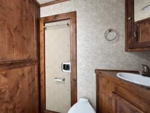 A small bathroom in the Logan Stock Combo Living Quarters features wood cabinets, a beige door with a toilet paper holder, towel ring, toilet, and a sink set in a wooden vanity. The walls are finished with light textured wallpaper. -Stillwater Trailer Sales