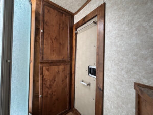A small Logan Stock Combo Living Quarters room with a wooden cabinet on the left wall and a beige door with a handle, lock, and metal rod above. The textured light beige walls are brightened by sunlight filtering through a frosted glass window. -Stillwater Trailer Sales