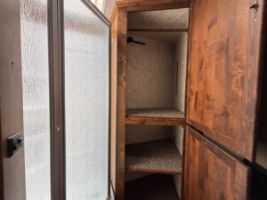 Open wooden closet in the Logan Stock Combo Living Quarters features two carpeted shelves beside a frosted glass shower door. Upper shelf includes a hanger, and walls have a beige finish. Closet door is partially open. -Stillwater Trailer Sales