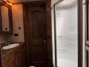 A bathroom inside the Logan Stock Trailer features a wooden door, small sink with cabinet and mirror, and a frosted glass shower enclosure. Warm lighting gives these combo living quarters a cozy feel. -Stillwater Trailer Sales