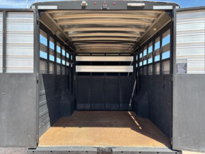Open view into the empty interior of a Logan Stock Combo trailer with metal walls, slatted side panels for ventilation, and a textured floor, seen from the rear with doors open under a sunny sky&mdash;ready for custom Living Quarters. -Stillwater Trailer Sales
