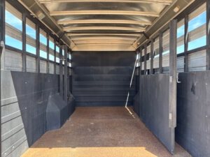 Interior view of an empty Logan Stock Combo livestock trailer with metal walls, a curved roof, and a textured brown floor. Sunlight filters through the side slats, creating patterns on the floor and walls. -Stillwater Trailer Sales