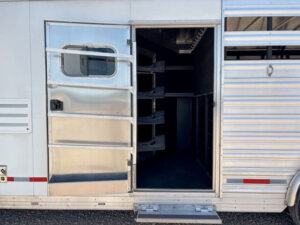 Open side door of a silver Logan Stock trailer showing an interior with multiple saddle racks and storage compartments. The door has a small window and an exterior step below it, perfect for Combo Living Quarters setups. -Stillwater Trailer Sales