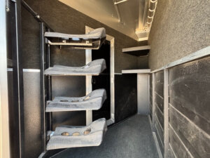 Interior of a Logan Stock Trailer featuring three molded saddle racks mounted vertically on the left wall, carpeted flooring and walls, and black storage shelves in the back. -Stillwater Trailer Sales