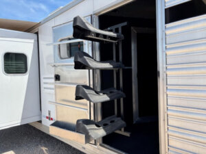 A close-up of a Logan Stock Combo Living Quarters horse trailer shows three black saddle racks mounted on the open tack room door, extending outward. The silver exterior features horizontal lines and a white door visible on the left. -Stillwater Trailer Sales