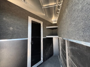 Interior of a small, narrow Logan Stock Combo trailer with gray carpeted walls, a black door with a metallic frame, and two wall-mounted shelves on the right. The Combo Living Quarters space is empty and well-lit. -Stillwater Trailer Sales