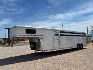 An Elite 28' Stock Trailer, featuring multiple side vents and dual axles, is parked on a gravel lot under a blue sky with some clouds. -Stillwater Trailer Sales