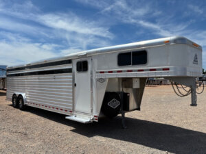 A large silver 28' Elite Livestock Trailer with multiple windows and dual axles is parked on a gravel lot under a blue sky with wispy clouds. -Stillwater Trailer Sales