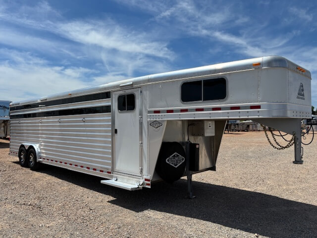 A large silver Elite Stock Trailer with multiple windows is parked on gravel under a blue sky with wispy clouds. -Stillwater Trailer Sales