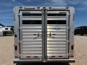 A close-up view of the rear of a silver Elite stock trailer with two closed, ribbed metal doors and red reflectors, parked on gravel under a clear blue sky. -Stillwater Trailer Sales