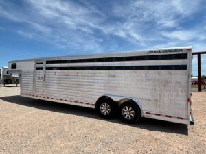 A large, silver 28' stock trailer with horizontal slats and dual axles is parked on gravel under a blue sky. The Elite trailer features black-trimmed windows along the upper side and shiny, multi-spoke wheels. -Stillwater Trailer Sales