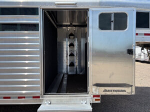 Open compartment of an Elite 28' Stock Trailer shows an organized tack room with three saddle racks mounted on the back wall and a fold-down step below the open door. -Stillwater Trailer Sales