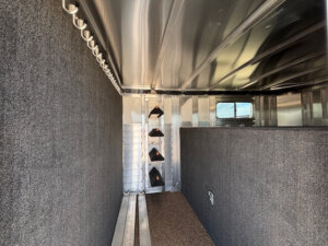 Interior of an Elite Stock Trailer with carpeted walls, hooks along the left side, a saddle rack holding three saddles near the center, and a window on the right. The non-slip floor adds safety for your horses and gear. -Stillwater Trailer Sales