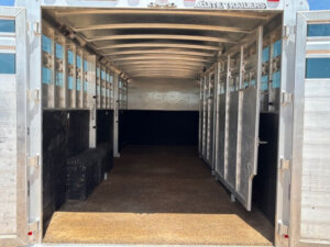 The image shows the interior of an empty Elite 28' Stock Trailer with metal walls, an open partition, and textured flooring. The ceiling is curved, and bright daylight comes from the open back doors. -Stillwater Trailer Sales
