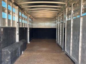The image shows the empty interior of an Elite Livestock Trailer with metal walls, a textured floor, and a curved ceiling. Sunlight filters through small side windows near the roof. -Stillwater Trailer Sales