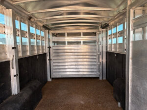 Interior view of an empty Elite Stock Trailer with ribbed walls, ventilation slats along the sides, and a closed gate at the far end. The textured floor and upper windows allow light to stream into this spacious livestock trailer. -Stillwater Trailer Sales