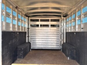 Interior view of an empty 28' stock trailer with metal walls, ventilation slats, nonslip flooring, and a central divider gate. Two black padded benches are on each side near the entrance. -Stillwater Trailer Sales