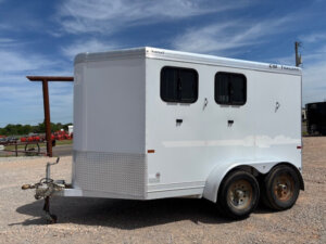 A white, two-axle CM Trailer 2 Horse Trailer is parked on gravel under a blue sky. The bumper pull trailer features two small windows with bars, a diamond plate on the front lower panel, and rust is visible on the wheels. -Stillwater Trailer Sales