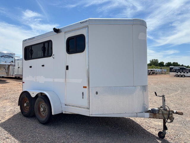 A white CM Trailers two-axle bumper pull trailer with black-tinted windows is parked on gravel under a blue sky with some clouds; other 2 horse trailers are visible in the background. -Stillwater Trailer Sales