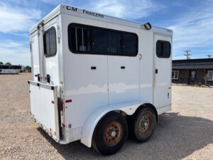 A white CM 2 Horse bumper pull trailer with small windows and rusted wheels is parked on a gravel lot under a blue sky. The trailer has "CM Trailers" written on the top edge. -Stillwater Trailer Sales