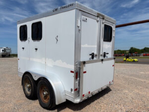 A white CM 2 Horse Trailer with two windows and dual rear wheels is parked on gravel under a blue sky. This bumper pull trailer shows some wear and rust on the wheels, with a yellow sports car and other vehicles in the background. -Stillwater Trailer Sales
