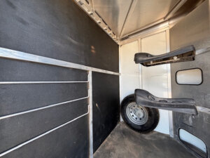 Interior of a CM horse trailer with black partition panels, saddle racks mounted on the wall, a spare tire on the floor, and metal rails along the side. This bumper pull model has a dusty floor. -Stillwater Trailer Sales
