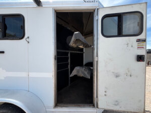 Open door of a CM 2 horse trailer reveals a dark interior with two saddle racks and metal railings; sunlight and gravel are visible outside this bumper pull. -Stillwater Trailer Sales