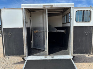 The image shows the open rear doors of a 2 Horse Bumper Pull CM Horse Trailer with a ramp down, revealing an empty, divided interior with metal partitions and barred windows. The trailer sits on gravel under a blue sky. -Stillwater Trailer Sales