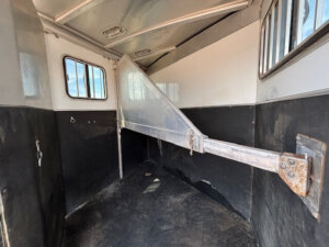 The interior of this clean and well-maintained 2 Horse Trailer features black and white walls, a central divider bar, side windows, and textured flooring&mdash;perfect for safe and comfortable travel. -Stillwater Trailer Sales