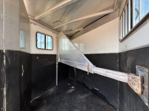 Interior of an empty CM 2 Horse Trailer with black and white walls, rubber flooring, a divider bar, and windows letting in natural light. This bumper pull design offers practical space and comfort for your horses. -Stillwater Trailer Sales