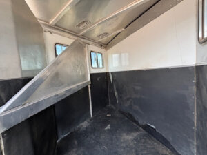 Interior of an empty CM 2 horse trailer with black and white walls, rubber flooring, metal dividers, and windows letting in natural light. The bumper pull design provides a clean, well-maintained space for safe equine transport. -Stillwater Trailer Sales