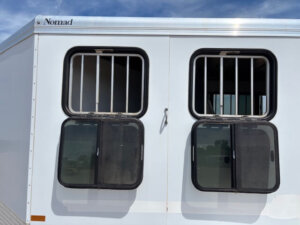 White CM Trailers bumper pull trailer labeled &ldquo;Nomad&rdquo; with two barred rectangular windows, each with two open black mesh screens, reflecting the blue sky and clouds above. -Stillwater Trailer Sales