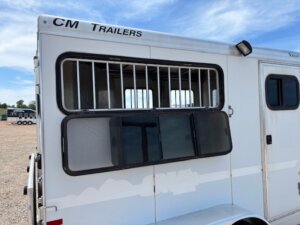 White CM 2 Horse bumper pull trailer with barred upper window and sliding lower window, parked on gravel. Another horse trailer is visible in the background under a blue sky with clouds. -Stillwater Trailer Sales