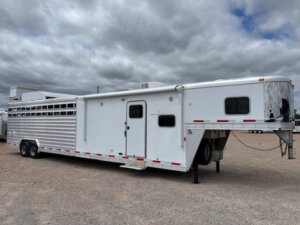 An Exiss Stock Combo livestock trailer with living quarters is parked on gravel under a cloudy sky. The large white trailer features windows, vents, and multiple axles for convenience and comfort on the road. -Stillwater Trailer Sales