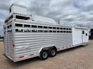 A large silver Exiss stock combo trailer with living quarters, ventilation slats, and dual axles is parked on gravel under a cloudy sky. The trailer features an air conditioning unit and a separate white door on the right side. -Stillwater Trailer Sales