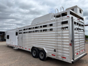 A large silver Exiss stock combo livestock trailer with air vents and a tall cooling unit on top is parked on a gravel lot under a cloudy sky. The trailer features tandem wheels and horizontal slats along its length. -Stillwater Trailer Sales